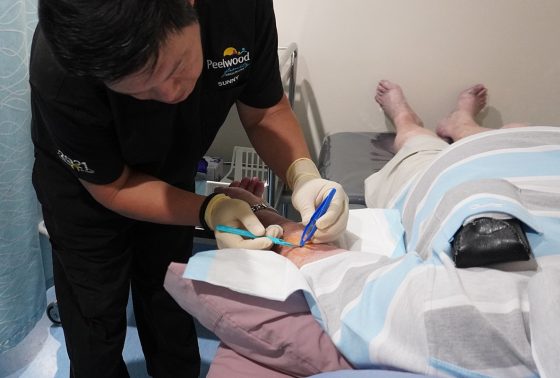 doctor with Peelwood Family Medical Practice scrubs on completing a minor surgery on a man's arm as he lays on a doctor's bed